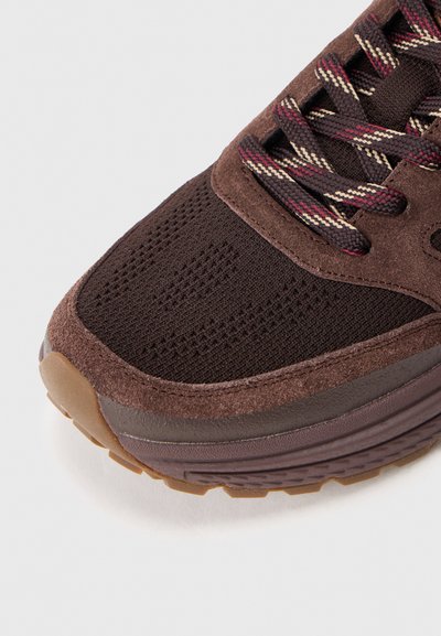 Vârf de sneaker maro cu material din suede și plasă, șireturi cu model marou și bej, și o talpă din cauciuc groasă, pe un fundal alb.