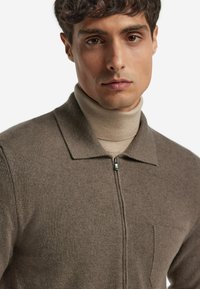 Brun dragkedjeströja med krage, tillverkad av ett mjukt tyg. Har en enda bröstficka och en texturerad stickad mönster. Modellen har en beige polotröja under.