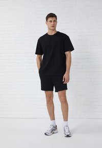 Zwarte T-shirt met korte mouwen en bijpassende shorts van zachte stof. Model draagt zilveren sneakers met witte sokken, staand voor een witte bakstenen achtergrond.