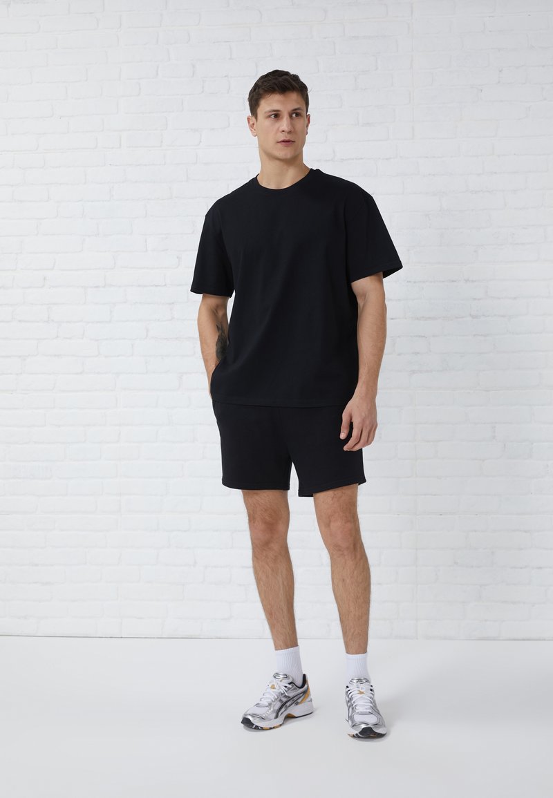 Zwarte T-shirt met korte mouwen en bijpassende shorts van zachte stof. Model draagt zilveren sneakers met witte sokken, staand voor een witte bakstenen achtergrond.