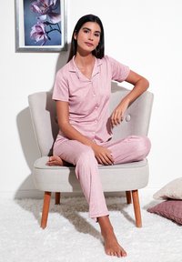 Roze pidžama s kratkim rukavima i uzorkom s točkicama, izrađena od mekog materijala. Model sjedi na svijetlosivoj stolici nasuprot bijelom zidu.
