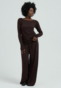 Woman standing wearing dark brown long-sleeve top and wide-leg pants, looking to the side against a plain light background.