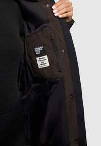 Sort frakke med en mørkebrun indvendig. Bemærkelsesværdige detaljer inkluderer en lynlås-lomme og stofetiketter, der angiver mærke og besked. Glat tekstur.