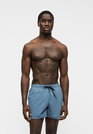 Homme torse nu à la peau foncée portant un short de bain bleu clair avec un cordon noir, debout devant un fond clair uni.