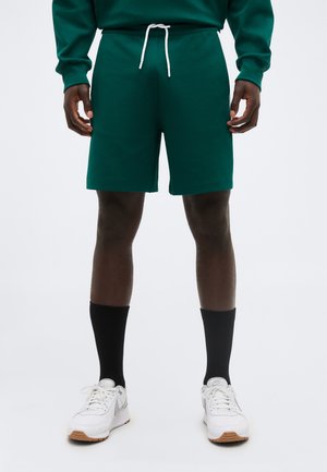 Person iført mørkegrønne shorts med hvid snor, sorte sokker til midt på læggen og hvide sneakers, stående mod en ensfarvet baggrund.