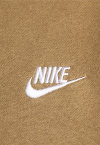 Beige tyg med en vit broderad Nike-logotyp och swoosh-design, som visar en slät, strukturerad yta.