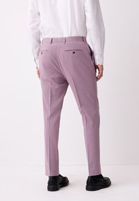 Liliowe spodnie garniturowe wykonane z gładkiej tkaniny, o dopasowanym kroju, z dwoma tylnymi kieszeniami i ciemnymi guzikami. Noszone z białą koszulą i czarnymi butami.