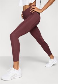 Maroon athletic leggings with a high waistband, featuring mesh panels and stitch detailing, paired with white sneakers.