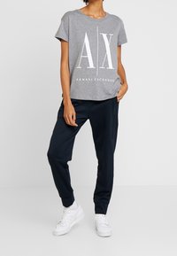 Šedé tričko s krátkým rukávem s velkým bílým logem "AX" a textem "ARMANI EXCHANGE" pod ním, spárované s tmavými tepláky a bílými teniskami.