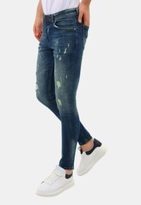 Slim-fit blauwe jeans met distressed details, rafelige stukken en een lichte wassing, gecombineerd met witte sneakers met zwarte accenten.