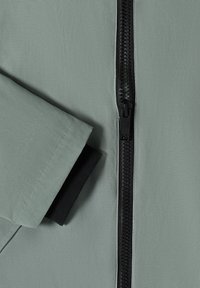 Manche de veste vert clair avec un poignet intérieur noir et une fermeture éclair en plastique noir centrée sur un tissu lisse.