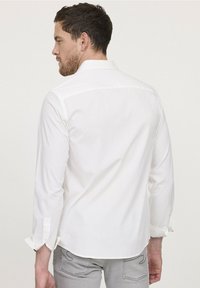 Chemise blanche à manches longues avec un col boutonné, un empiècement de dos droit et des poignets retroussés, portée par une personne de dos, sur un fond uni.
