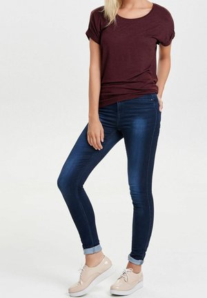 Frau trägt ein bordeauxrotes Kurzarmshirt, dunkelblaue enge Jeans, an den Knöcheln hochgekrempelt, und beige Schnürschuhe, steht vor weißem Hintergrund.