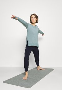 Sportoutfit mit einem hellblauen Oberteil mit seitlichen Schlitzen und dunklen, navyblauen Hosen. Auf einer grauen Yogamatte getragen, ist das Model barfuß in einer Pose.