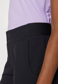 Černé kalhoty s hladkou strukturou mají elastický pas a dvě boční kapsy. Částečně je vidět světle fialový top.