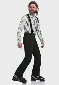 Schwarze Skihosen mit verstellbaren Trägern, kombiniert mit einem weißen, marmorierten Langarmshirt und schwarzen Skischuhen mit pinken Akzenten.