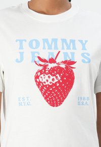 T-shirt en coton blanc avec un grand graphique de fraise rouge, texte bleu "TOMMY JEANS", et accents bleus pour "EST. N.Y.C." et "1985 U.S.A."