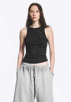 Vrouw met lang donker haar, gekleed in een zwart mouwloos topje met studs en lichtgrijze joggingbroek, handen in de zakken, tegen een witte achtergrond.