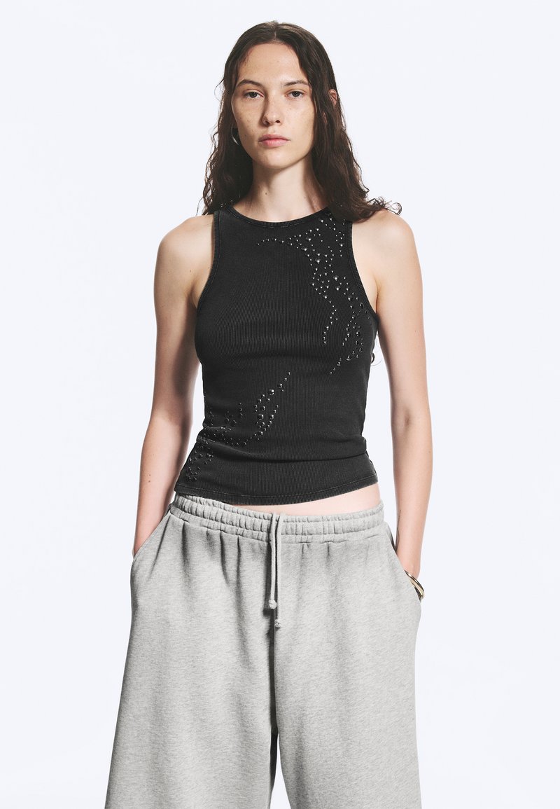 Femme aux longs cheveux foncés portant un haut noir sans manches avec des clous et un pantalon de survêtement gris clair, mains dans les poches, sur fond blanc.