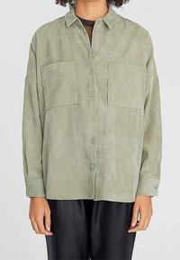 Persona che indossa una camicia verde chiaro con bottoni, dotata di due grandi tasche sul petto e pantaloni neri, in piedi contro uno sfondo semplice.