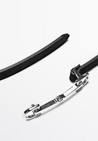 Čern�ý kožený řemínek s lesklou stříbrnou kovovou přezkou, obsahující T-tvarovaný designový prvek a hladkou texturu.