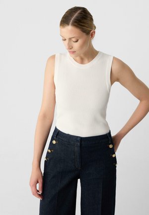 White ribbed tank top paired with dark denim wide-legged trousers featuring gold button accents on the sides. Smooth texture, fitted design.