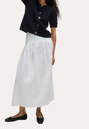 Femme portant un cardigan noir à manches courtes avec des boutons dorés, une jupe blanche longueur cheville et des ballerines noires.