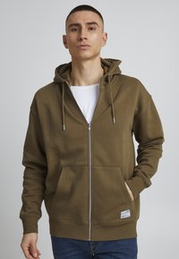 Olivgrüner Zip-Hoodie mit Kängurutasche, verstellbare Kapuze und gerippten Bündchen. Hergestellt aus weichem Stoff und mit einem Logosticker am Saum.