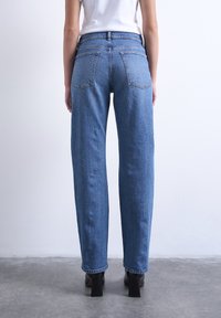 Άτομο που φοράει μπλε τζιν με ίσια γραμμή, λευκό πάνω μέρος και μαύρα μποτάκια με τακούνι, στέκεται σε γκρι πάτωμα μπροστά σε λευκό τοίχο.