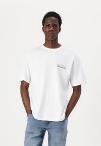 Hvid bomulds T-shirt med korte ærmer og rund halsudskæring, med et lille grønt "Cancun" grafisk motiv på venstre brystområde.