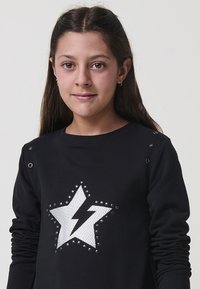 Mlada deklica z dolgimi rjavimi lasmi nosi črno jopico z belim motivom zvezde z obliko strele.