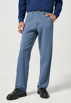 Jasnoniebieskie bawełniane spodnie o prostym kroju, z tylną kieszenią i zapięciem na guzik. Noszone z czarnymi butami, co podkreśla swobodny wygląd.