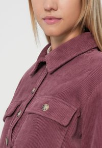 Πουκάμισο καπιτονέ σε ροζ σκόνη, με δύο μπροστινές τσέπες, γιακά που διπλώνει και κουμπιά. Υφή υφάσματος με κατακόρυφες ραβδώσεις.