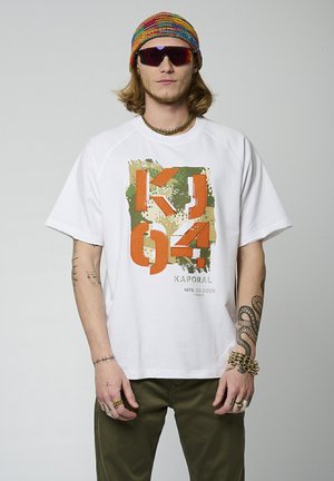 T-shirt en coton blanc avec un motif graphique en orange, vert et beige. Caractéristiques : coupe décontractée et manches raglan, montré avec un pantalon olive.