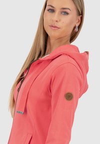 Korallenfarbener Zip-Hoodie mit einer strukturierten Kapuze, Seitentaschen und einem Logo-Patch am Ärmel. Verfügt über einen glatten Stoff und eine sportliche Passform.