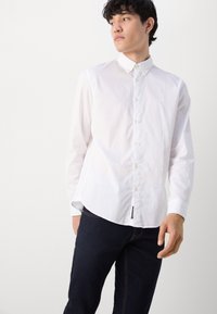 Weißes Button-Down-Hemd aus leichtem Baumwollstoff mit langen Ärmeln, abgerundetem Saum und einem kleinen Logodetail auf der Brust. Kombiniert mit dunkelblauen Jeans.