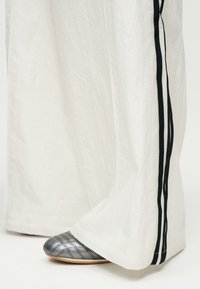 Λευκό παντελόνι με φαρδιά πόδια και διπλές μαύρες ρίγες στο πλευρό, που καλύπτει μερικώς ένα ασημί πλατφόρμα παπούτσι με ξύλινη σόλα και σχέδιο καπιτονέ.