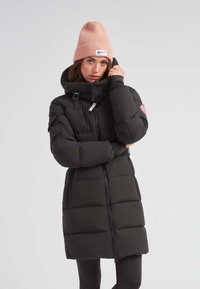Abrigo acolchado negro con cuello alto, cierre de cremallera y capucha desmontable, combinado con un suave gorro de punto rosa que cuenta con una etiqueta.