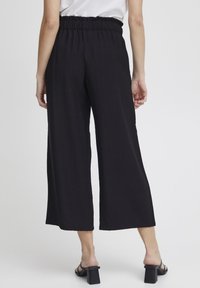 Zwarte, wijde broek met een elastische tailleband en een gestructureerde stof. De broek is enkel-lang en is gecombineerd met zwarte hakken.