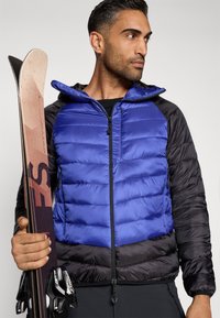 Casaco puffer azul e preto com design acolchoado, zíperes frontais e capuz. Modelo está a segurar esquis, apresentando um padrão texturizado e acentos escuros.