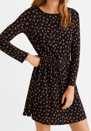 Femme portant une robe noire à manches longues avec un petit motif floral multicolore et une taille cintrée avec un cordon de serrage.