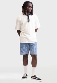 Cremefarbiges Oversized-T-Shirt, blaue Jeansshorts mit Flammenmuster, schwarze runde Sonnenbrille, schwarze Schuhe und weiße Socken.