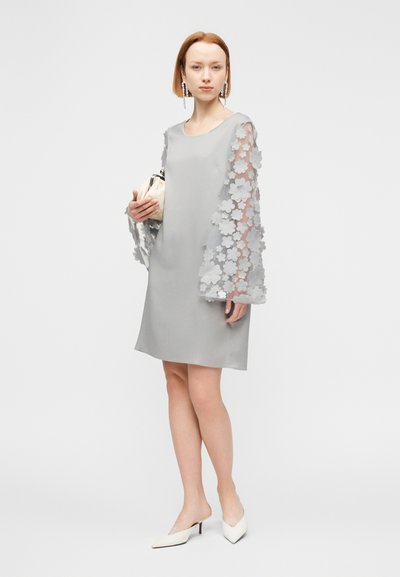 Γκρι φόρεμα με διαφανή μανίκια από δαντέλα με λουλούδια, λεία υφή, άνετη εφαρμογή. Το μοντέλο κρατά μια μικρή κρεμ τσάντα χειρός και φορά μυτερές λευκές γόβες.