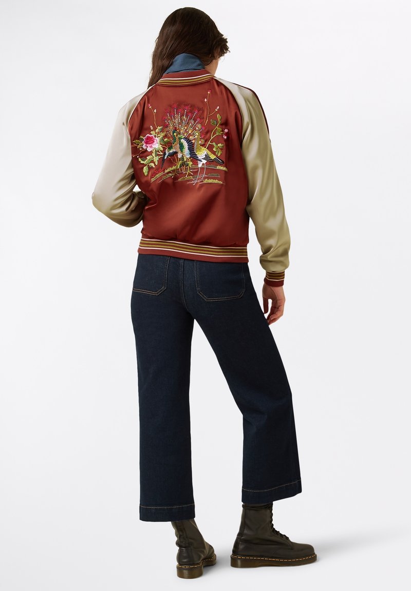 Red and beige bomber jacket with embroidered floral and bird design; ribbed cuffs and hem; paired with dark denim wide-leg jeans and black boots.