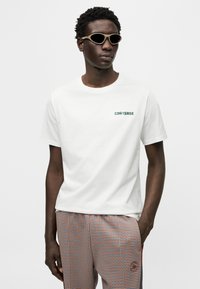 Λευκό βαμβακερό t-shirt με πράσινο λογότυπο "CONVERSE", σε συνδυασμό με παντελόνι με πορτοκαλί και μπλε καρό σχέδιο. Το μοντέλο φορά σκοτεινά γυαλιά ηλίου.