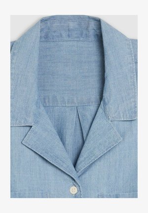 Chemise chambray bleu clair avec un col à encoches, une fermeture à un bouton et une texture subtile avec des détails de couture minimalistes.