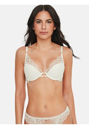 Femme portant un soutien-gorge en dentelle blanche et une culotte en dentelle assortie, debout devant un fond clair uni, faisant face à l’avant.