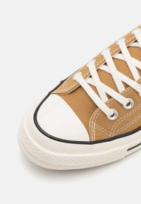 Zapatillas de lona marrones con puntera y suela de goma blanca, que cuentan con cordones blancos y acentos de ojales, costuras detalladas y ribete negro.