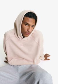 Világos rózsaszín kapucnis pulóver elülső zsebbel és hímzett logóval; világosszürke sweatpantsszel párosítva. Sima textúra, laza szabás, minimalista dizájn.