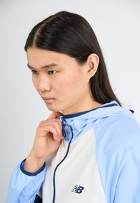 Ľahká bunda s modro-bielou farebnou kombináciou. Obsahuje zips na golieri a logo New Balance na prednej ľavej strane. Hladká textúra.